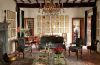 a room with a crystal chandelier, vintage sofa and chairs with velvet cushions, cocktail table with candelabras and trays, oriental rug, wooden desk and chair, a wall with 21 small framed botanical art pieces, beamed ceiling
