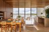 a large open room with a glass wall looking out to the sea, dining table and chairs, sofa, loveseat, and cocktail table, slatted wood wall with inset shelves, kitchen island with sink and cabinets, wood ceiling and floor