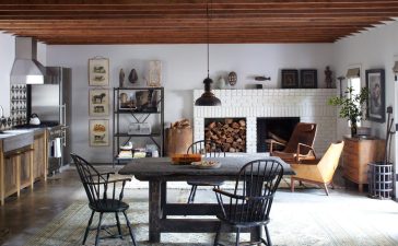 wood beam ceiling living dining space with a white brick fireplace at far end and a wood insert next to it and some modern wood club chairs and a dark square farmhouse table and chairs in the foreground and a kitchen set up to the left